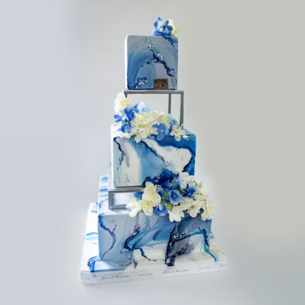 Scopri la Torta Sogno Marino di Gocce di Cioccolato, con tre piani di sfumature marine e decorazioni di fiori bianchi e blu, perfetta per eventi speciali.